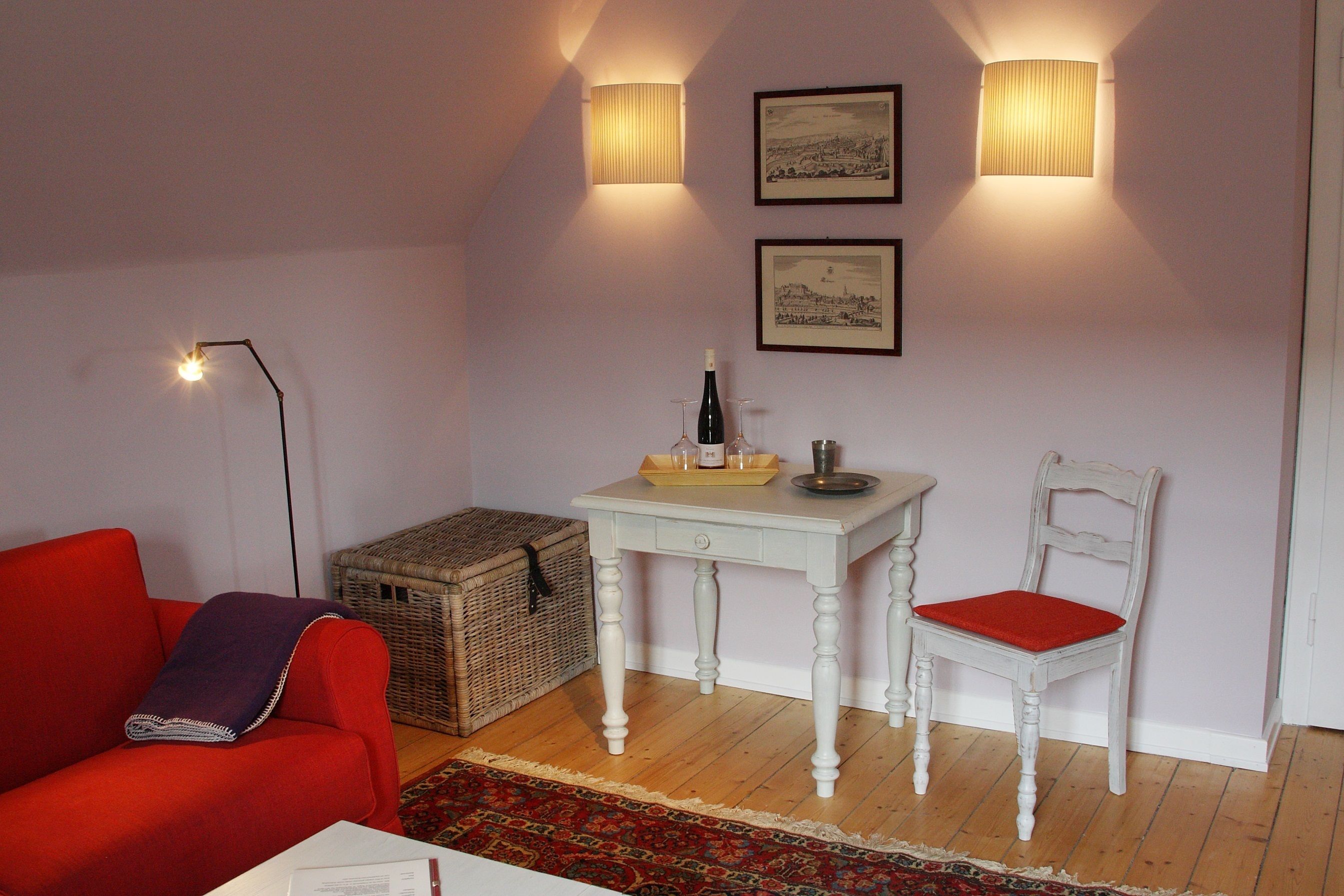 Una habitación acogedora con un sofá rojo, una mesa blanca con una botella y vasos, una cesta de mimbre y una silla blanca con un cojín rojo.