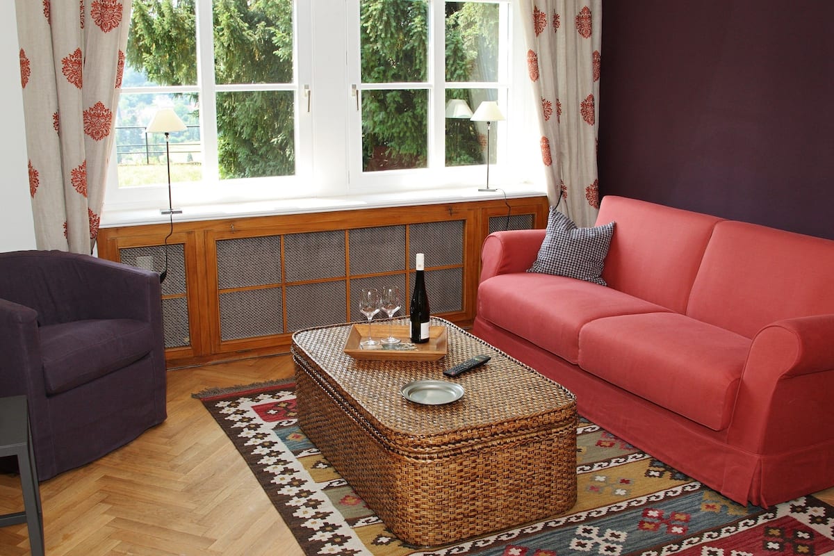 Un salón con un sofá rojo, un sillón púrpura, una mesa de centro de mimbre con una bandeja que sostiene una botella y vasos, y una alfombra estampada.