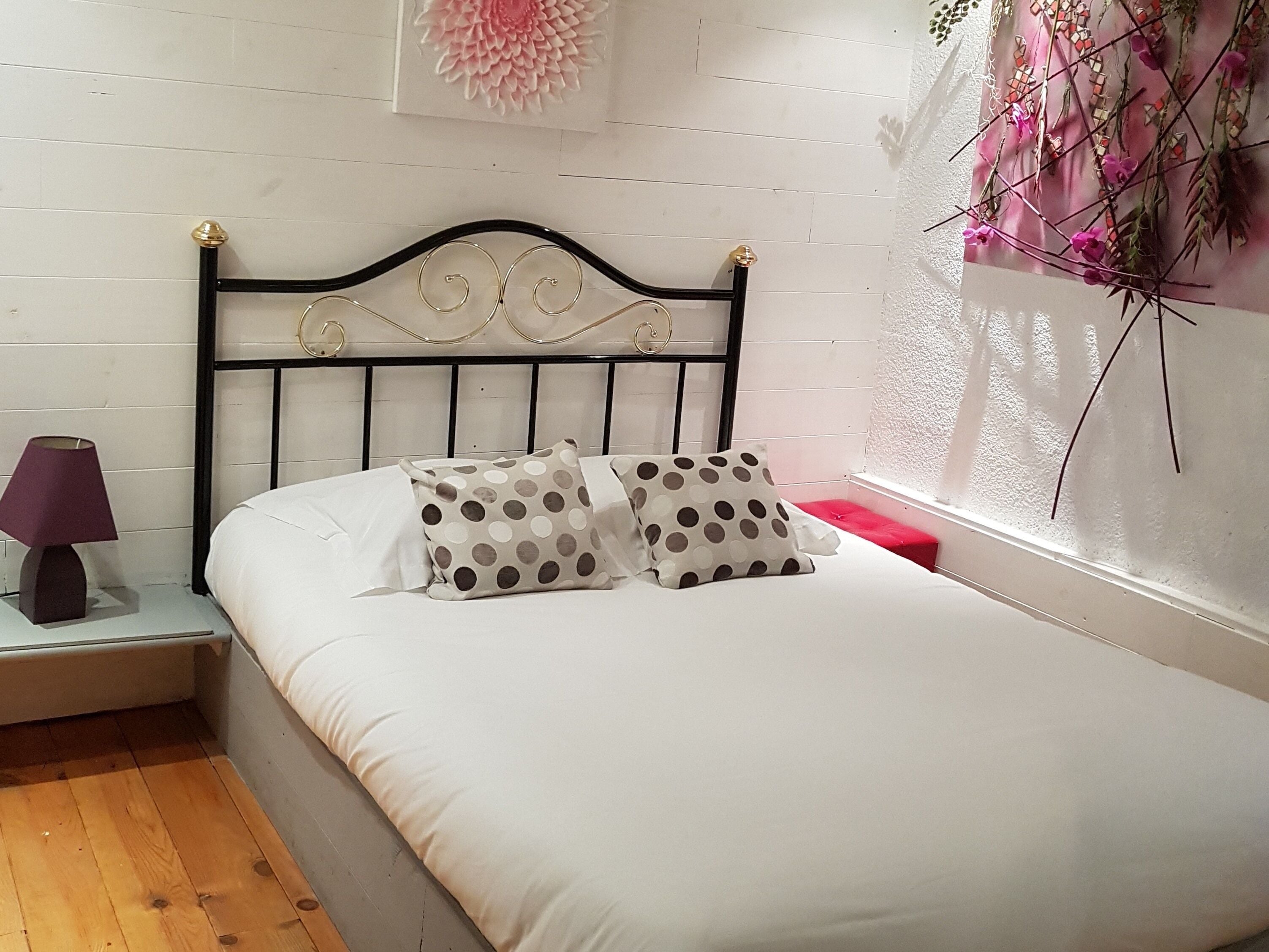 Een bed met hoofdeinde, twee kussens, een nachtkastje met lamp en een raam met plant.