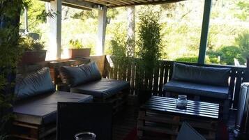 Un patio exterior con muebles de madera, incluyendo un sofá y una mesa, rodeado de plantas en macetas.