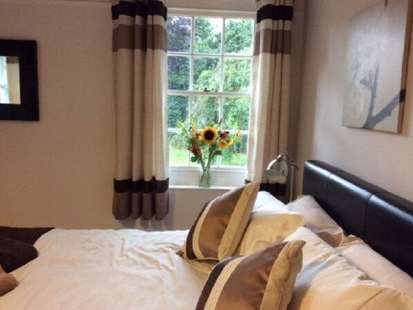 Un dormitorio con cama, cortinas, ventana con vista a follaje y un cuadro en la pared.