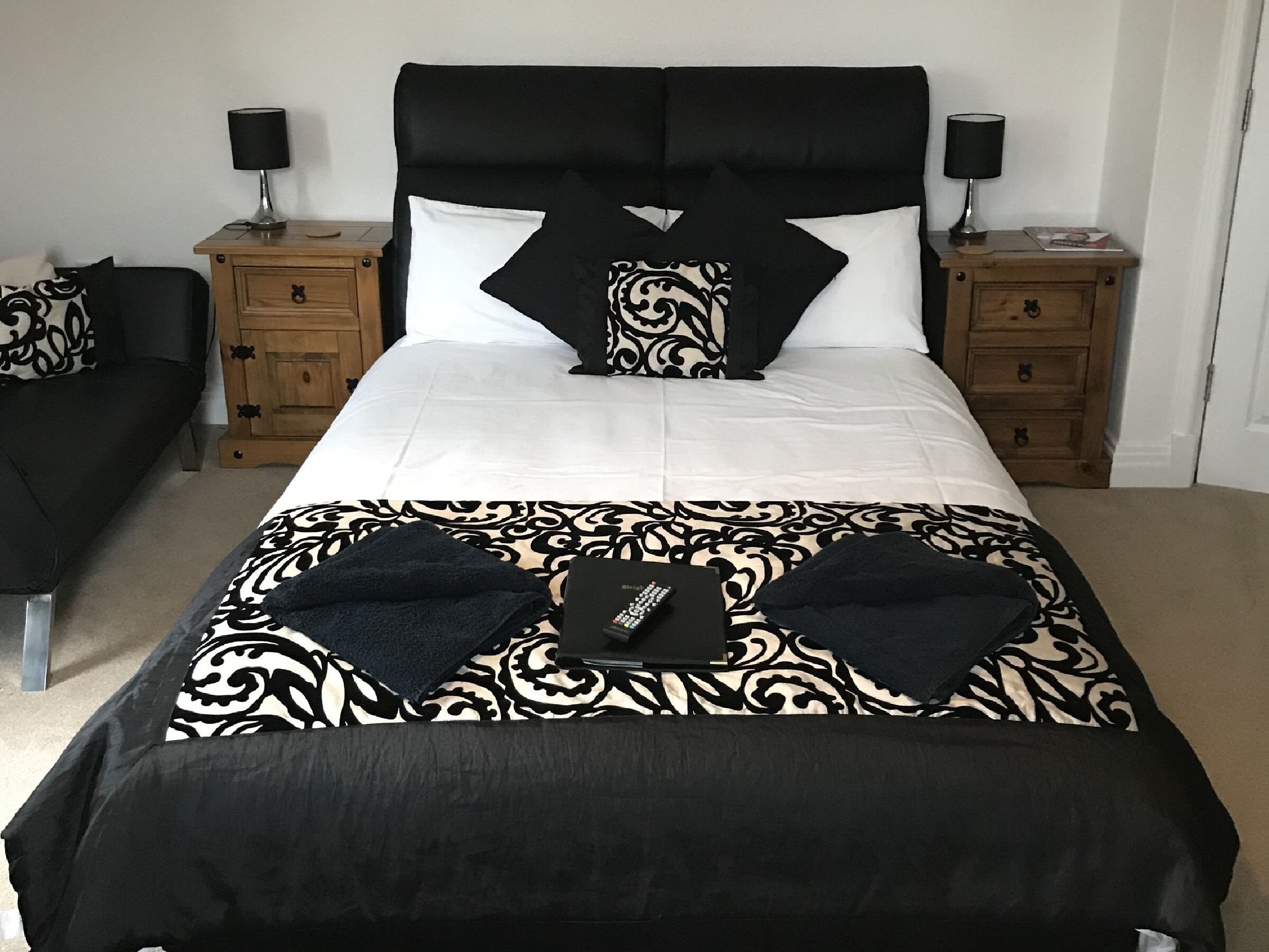 Une chambre à coucher avec un grand lit, deux tables de chevet, une petite table d’appoint, un canapé noir et un plaid à motifs.