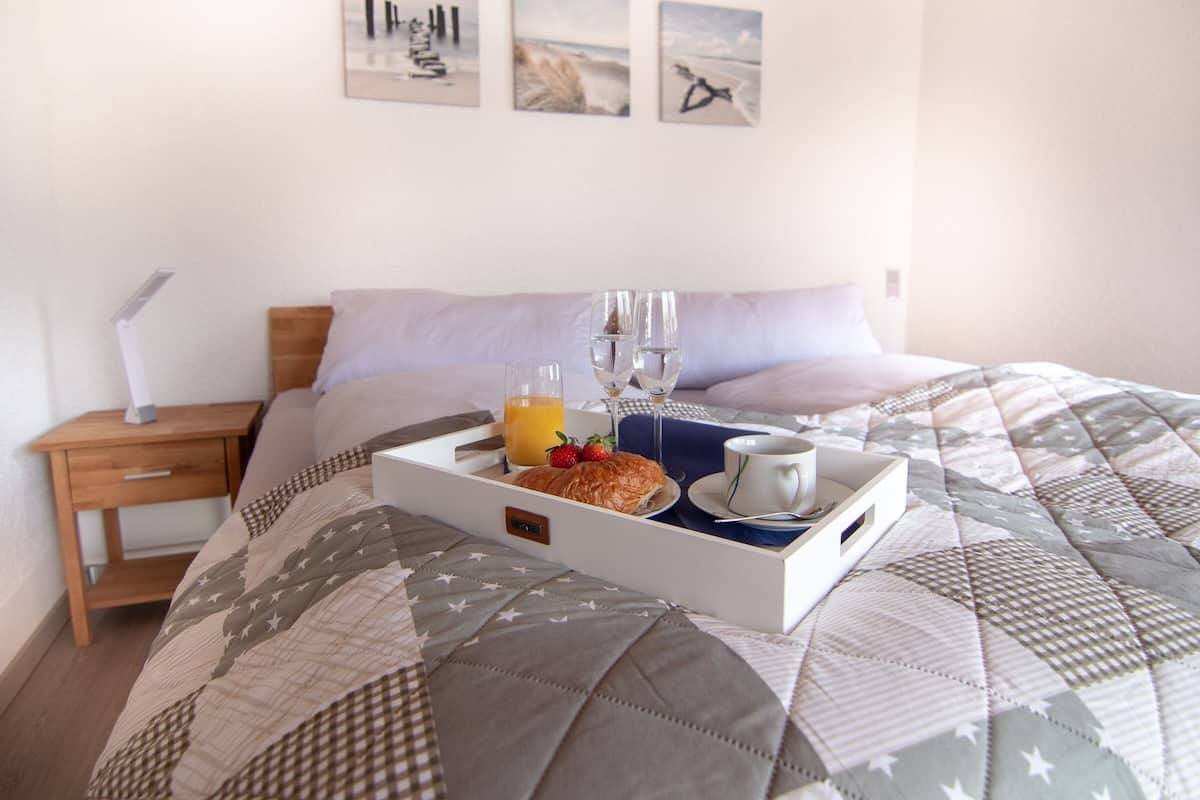 A neatly made bed with a quilt, a bedside table with a lamp, and a tray with breakfast items including croissants, juice, and wine glasses.