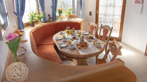 Ein Essbereich mit einem runden Tisch, gedeckt für das Frühstück, umgeben von Sitzgelegenheiten mit Kissen und großen Fenstern mit blauen Vorhängen.
