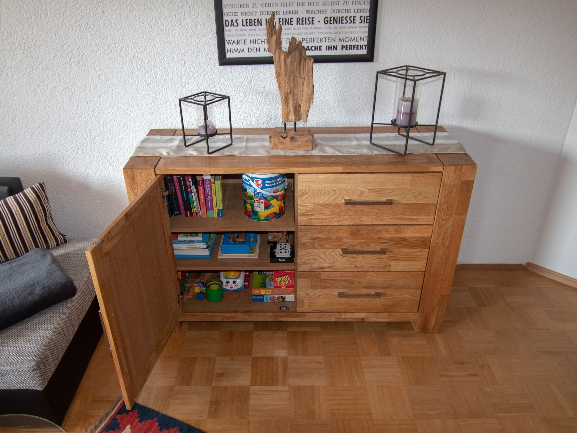 Un mueble de madera con estantes abiertos que contienen libros y juguetes, una cita enmarcada en la pared, y una escultura de madera encima.