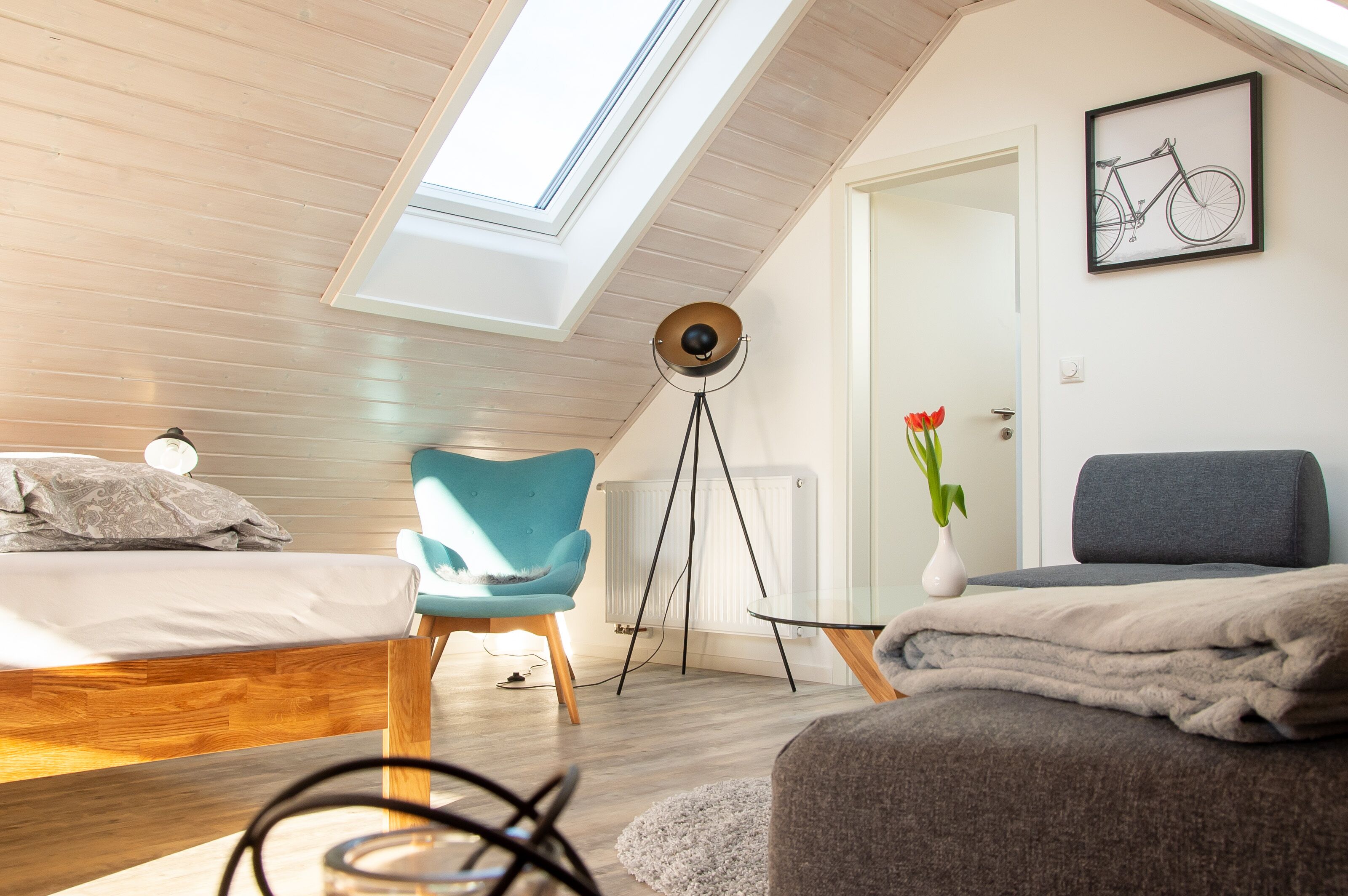 A modern bedroom with a skylight, a bed, a blue chair, a floor lamp, and a framed bicycle artwork.