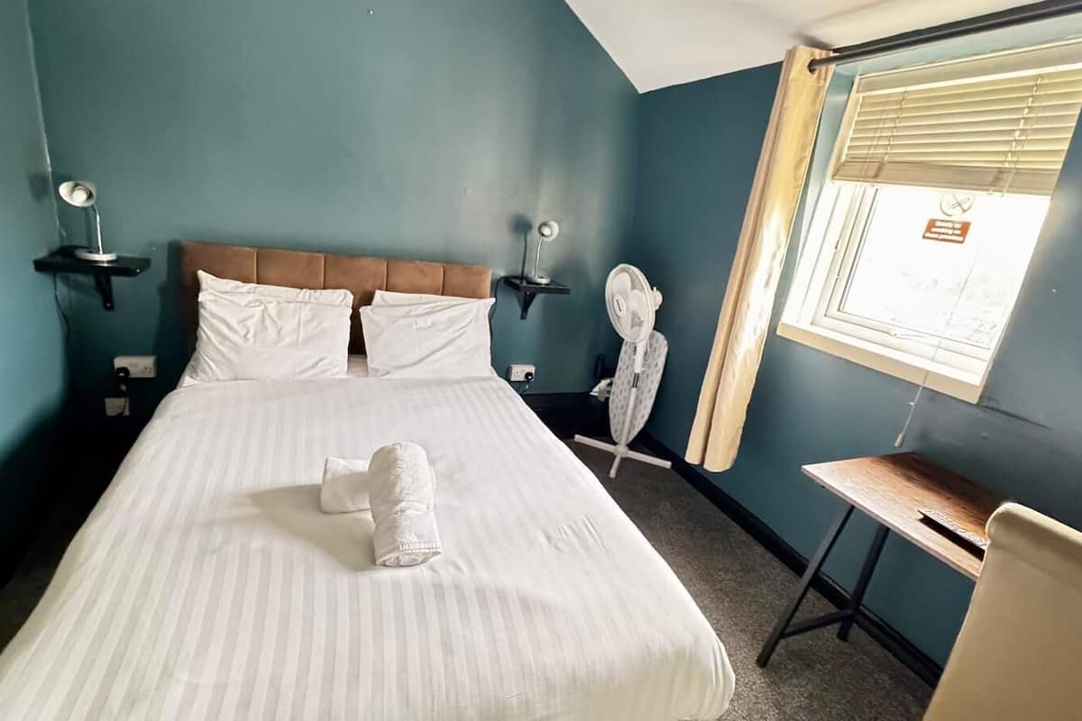 A hotel room with a large bed, two bedside tables with lamps, a desk, and a window with blinds.