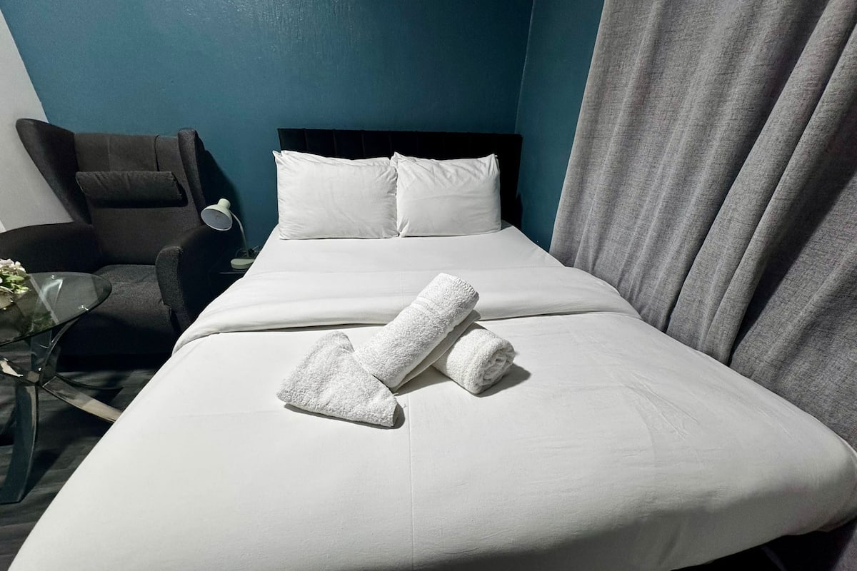 A neatly made bed with white linens and two rolled towels, a black headboard, a grey curtain, and a black armchair with a small table beside it.