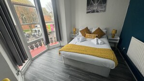 A hotel room with a bed, two nightstands, a window with curtains, a yellow throw blanket, and a painting on the wall. - Avalon Guest House (Brighton)