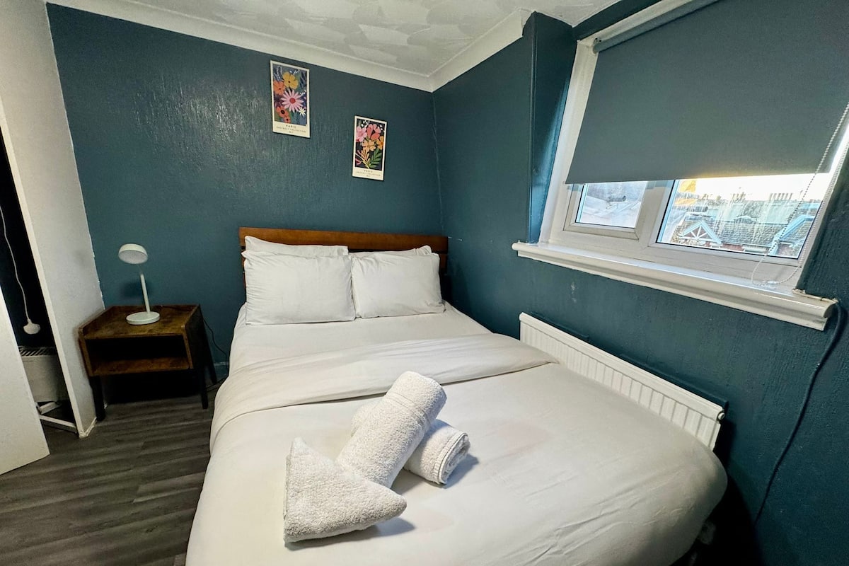 A neatly made bed with white linens and two towels, a wooden nightstand with a lamp, and a window with a view of buildings.