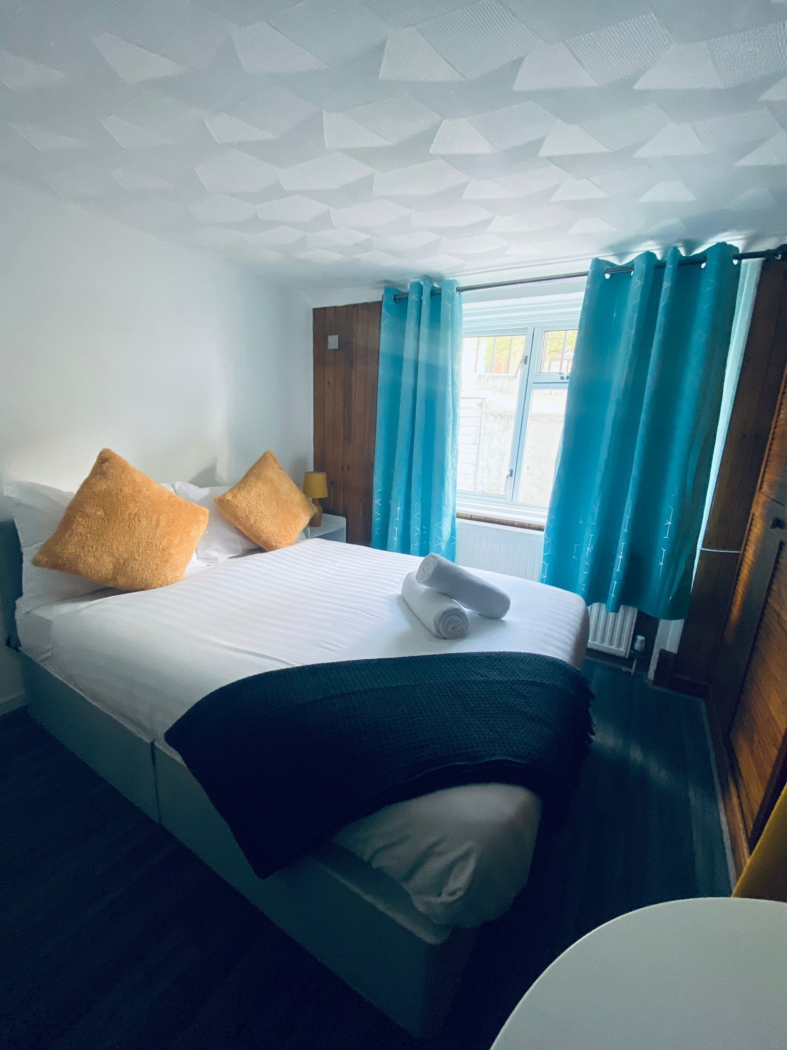 Une chambre d’hôtel avec un lit, des oreillers, une serviette roulée, une fenêtre avec des rideaux et une porte en bois.