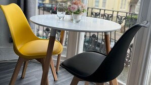 A room with a round table, two chairs, a vase with flowers, and a view of buildings through large windows. - Avalon Guest House (Brighton)