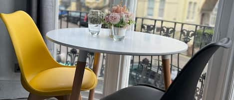 A room with a round table, two chairs, a vase with flowers, and a view of buildings through large windows.