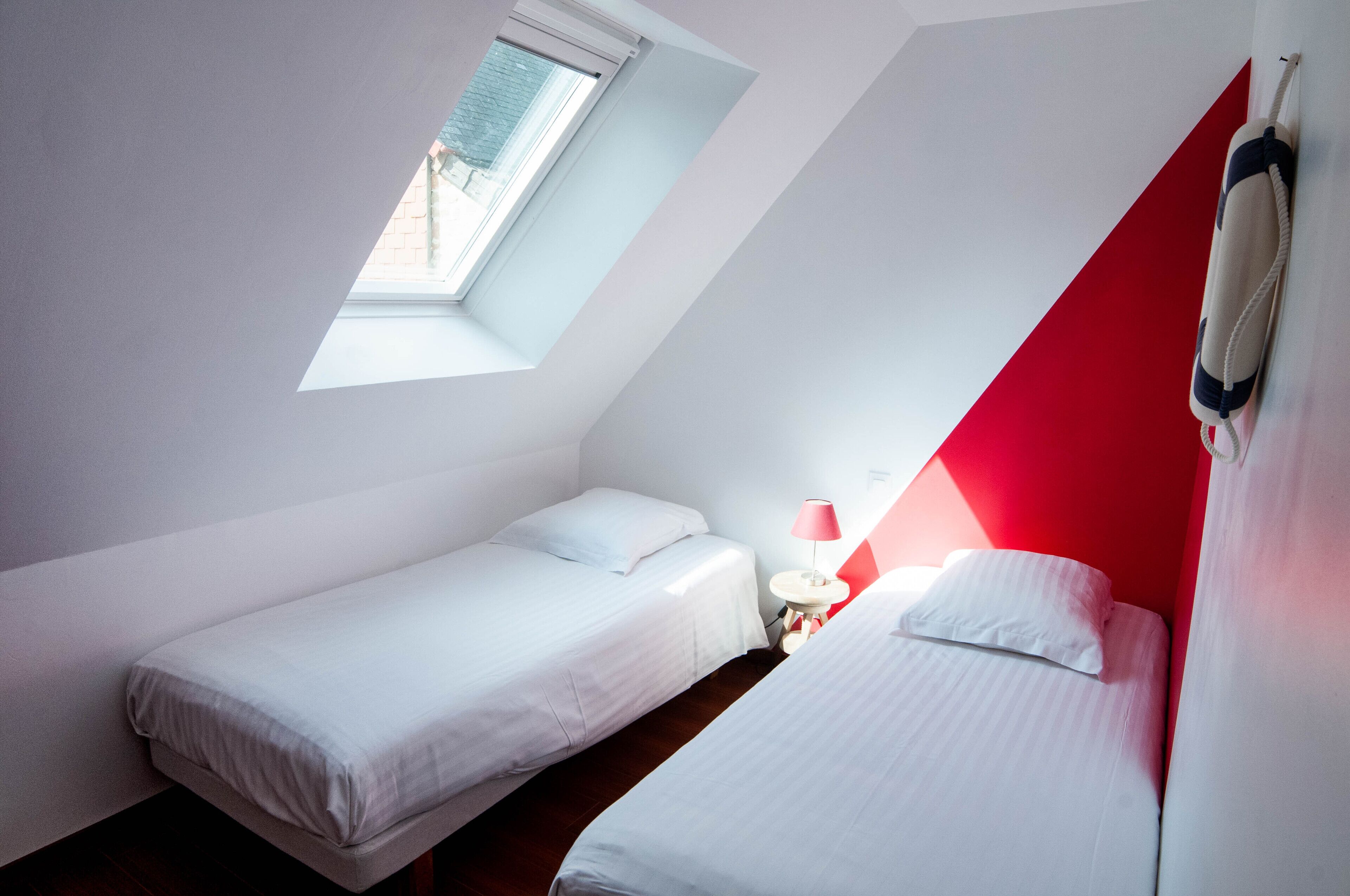 Une petite chambre moderne, dotée de deux lits simples, d’une petite table avec une lampe et d’une fenêtre laissant entrer la lumière naturelle.