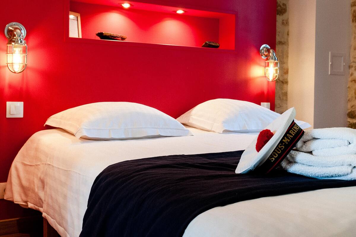 Een hotelkamer met een rode muur, een bed met wit beddengoed, twee wandlampen en een handdoek met een boek erop.