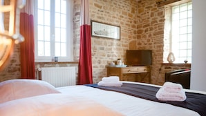 A bedroom with a stone wall, a bed with white linens, a TV, and a window with curtains. - La Ferme D'ervée de Saint-Roch (Isigny-sur-Mer)
