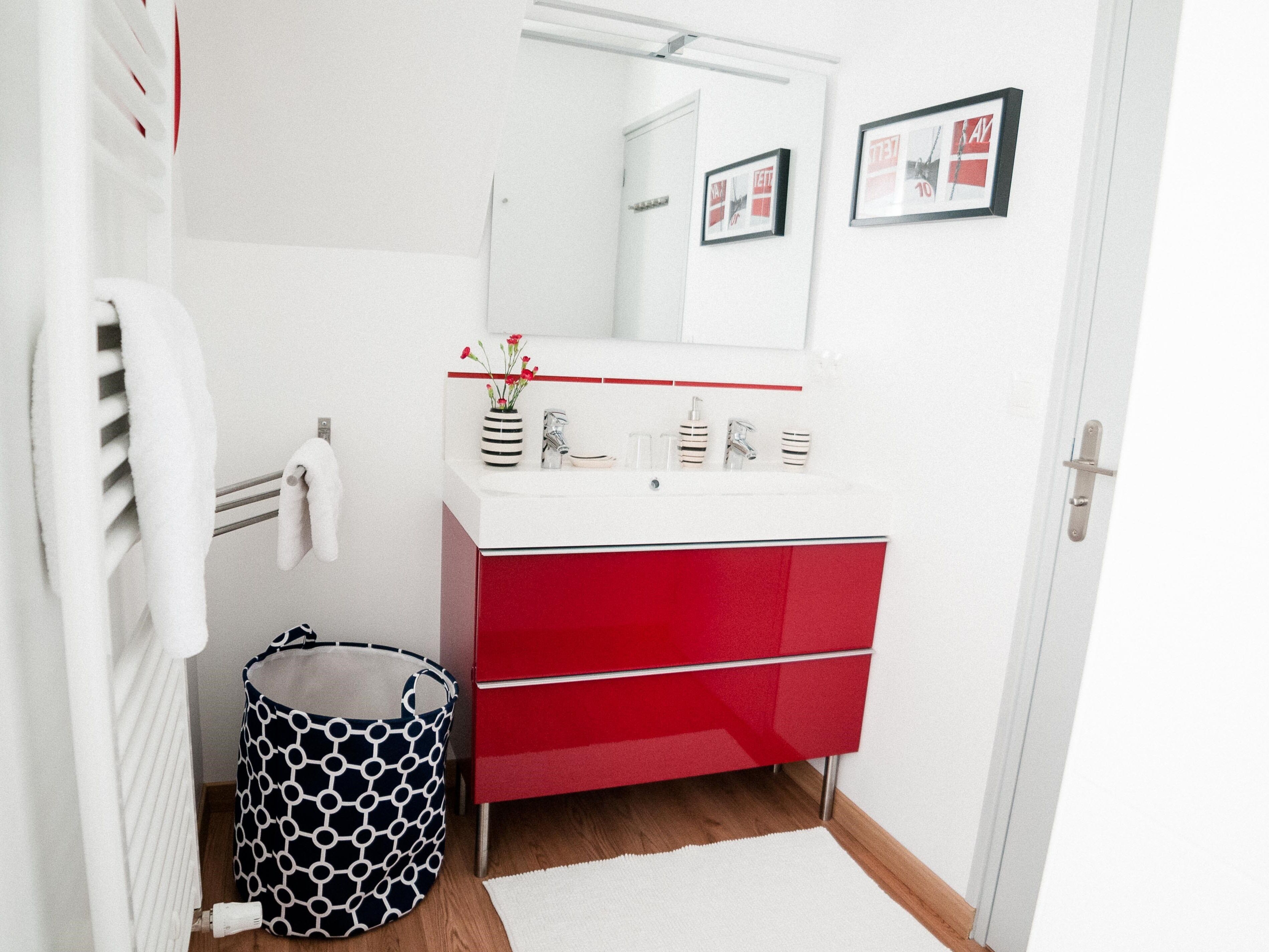 A modern bathroom with a white sink, red cabinet, and a mirror above it. There is a white towel hanging on a rack and a black and white patterned trash bin.