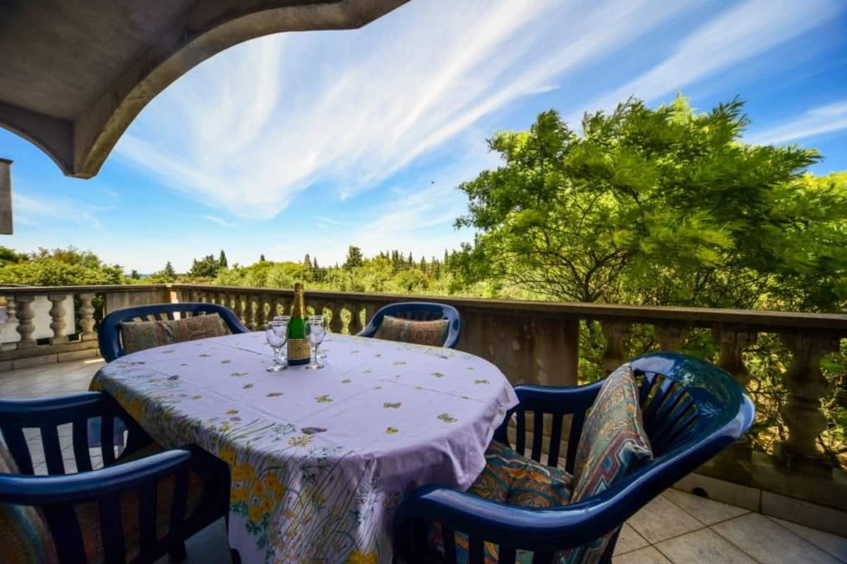 Une table ronde recouverte d’une nappe à motifs floraux, entourée de chaises bleues agrémentées de coussins à motifs, est placée sur un balcon d’où l’on peut admirer des arbres verdoyants et un ciel dégagé.