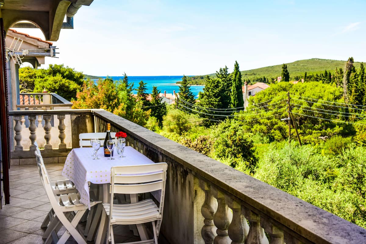Un balcon avec une table dressée pour deux personnes, offrant une vue sur un paysage côtier, avec des arbres et la mer à l’horizon.