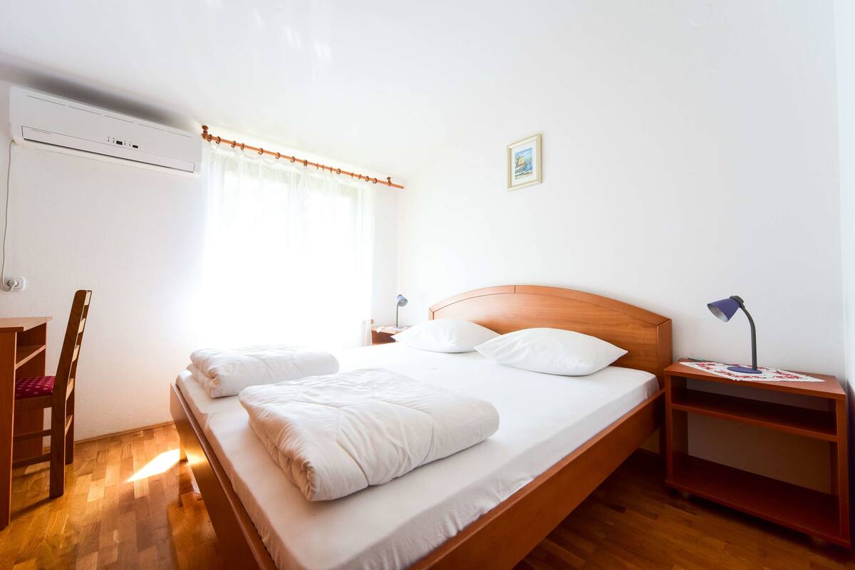 Un lit bien fait, avec du linge de lit blanc, une tête de lit en bois, une table de chevet avec une lampe et un petit bureau avec une chaise.