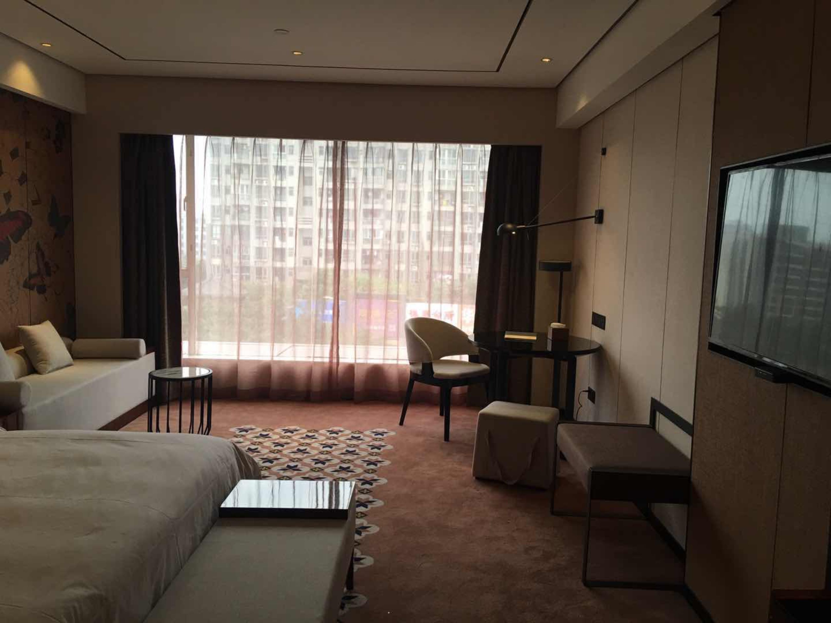 Et moderne hotelværelse med en stor seng, en sofa, et skrivebord og en stol. Der er et stort vindue med lette gardiner, et fladskærms-tv ophængt på væggen og et tæppe med et mønster på gulvet.