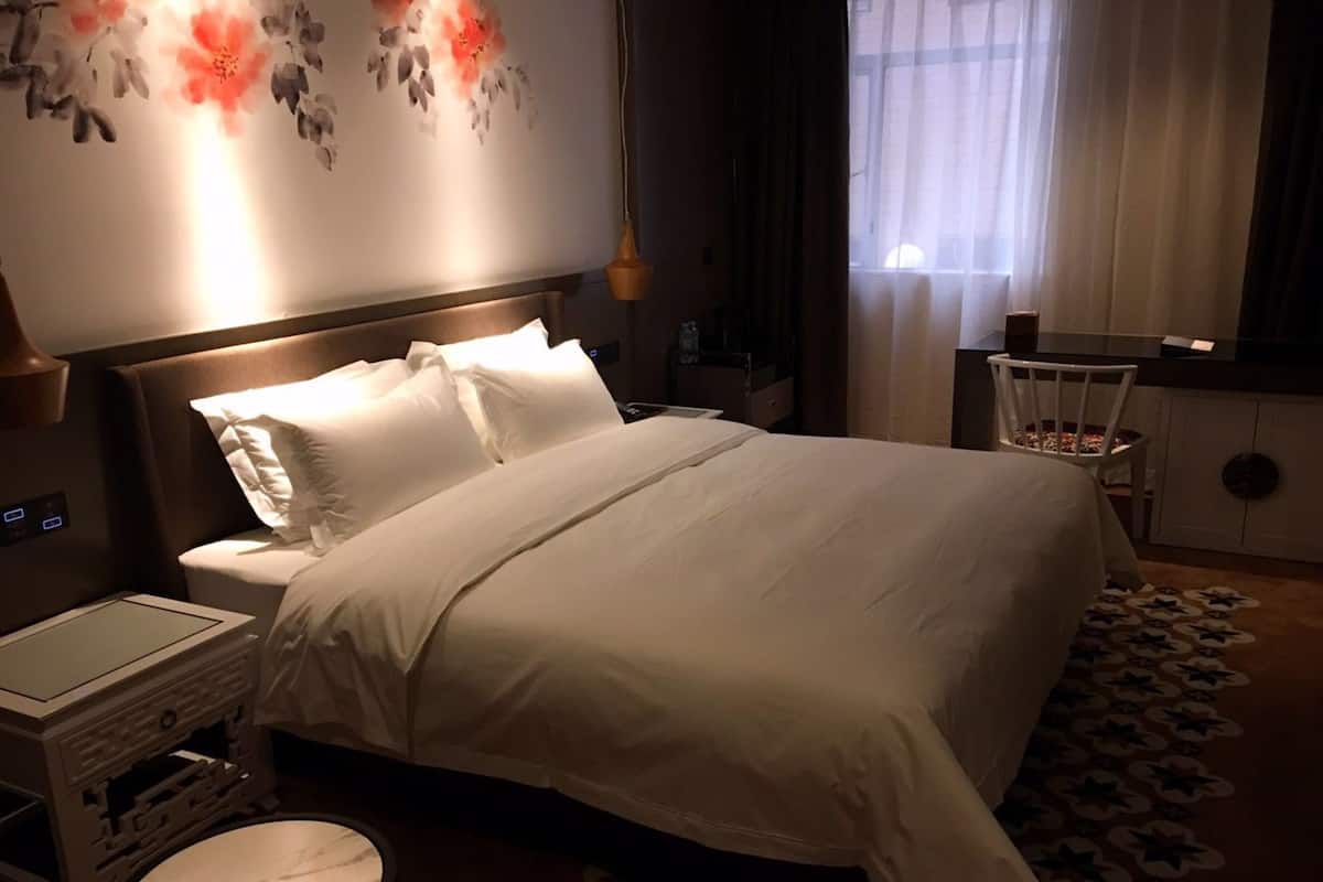 Habitación de hotel con una cama grande, una mesita de noche, una pequeña mesa redonda, una pared con motivos florales y una ventana con cortinas.