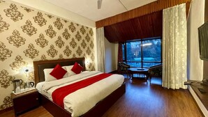 A hotel room with a large bed, a wooden nightstand, a desk, and a view of the outdoors through a large window. - Hotel Sitara international (Manali)