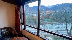 A room with a large window offering a view of a mountainous landscape and a river. - Hotel Sitara international (Manali)