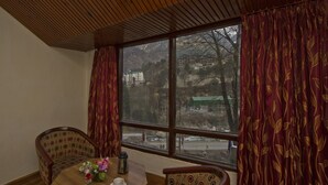 A dining area with a wooden table and chairs, a window with curtains, and a view of a mountainous landscape. - Hotel Sitara international (Manali)