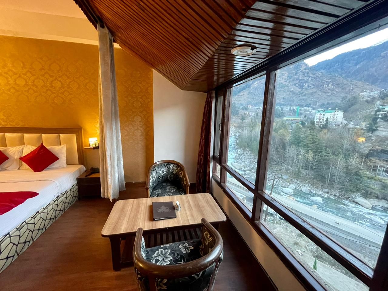 Ein Hotelzimmer mit Bett, Tisch und Stuhl, einem Fenster mit Blick auf einen Fluss und Berge, sowie einer Holzbalkendecke.