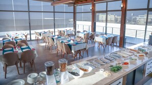 A spacious dining area with multiple tables and chairs, a buffet with various food items, and large windows offering a view of the city. - Carissa Hotel (Mugla)