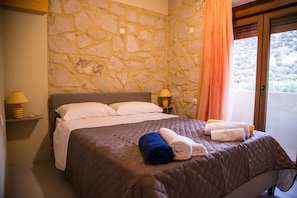 A bedroom with a stone wall, a bed with white and brown bedding, two bedside lamps, and a window with a curtain. - Uphoria Stone Villas (Mylopotamos)