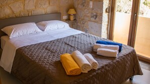 A bedroom with a stone wall, a bed with a quilted brown bedspread, white pillows, and rolled towels on the bed. - Uphoria Stone Villas (Mylopotamos)