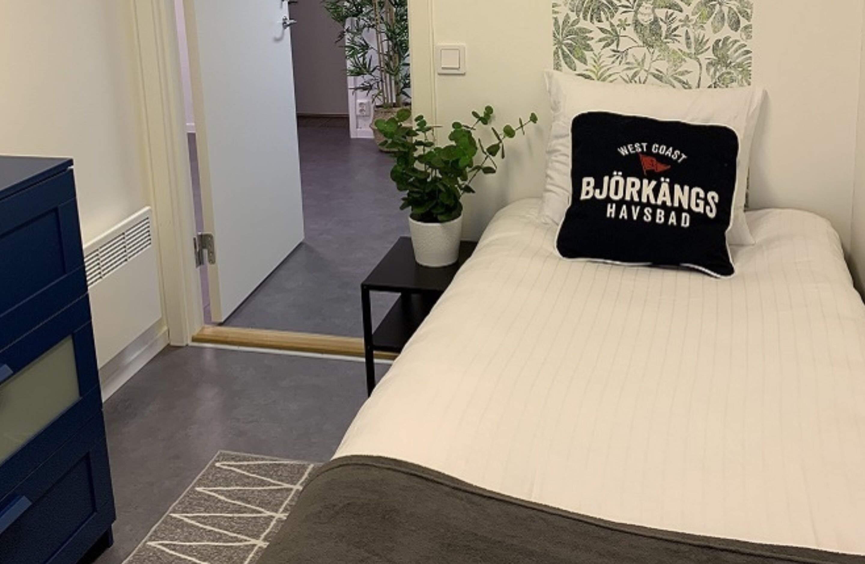 Une chambre à coucher avec un lit, une table de chevet ornée d’une plante, une commode bleue et une porte donnant sur une autre pièce.