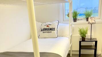 A bunk bed with a pillow featuring "West Coast Björkängs Håvåd" text, a small table with a plant, and a window with a view of a building and greenery.