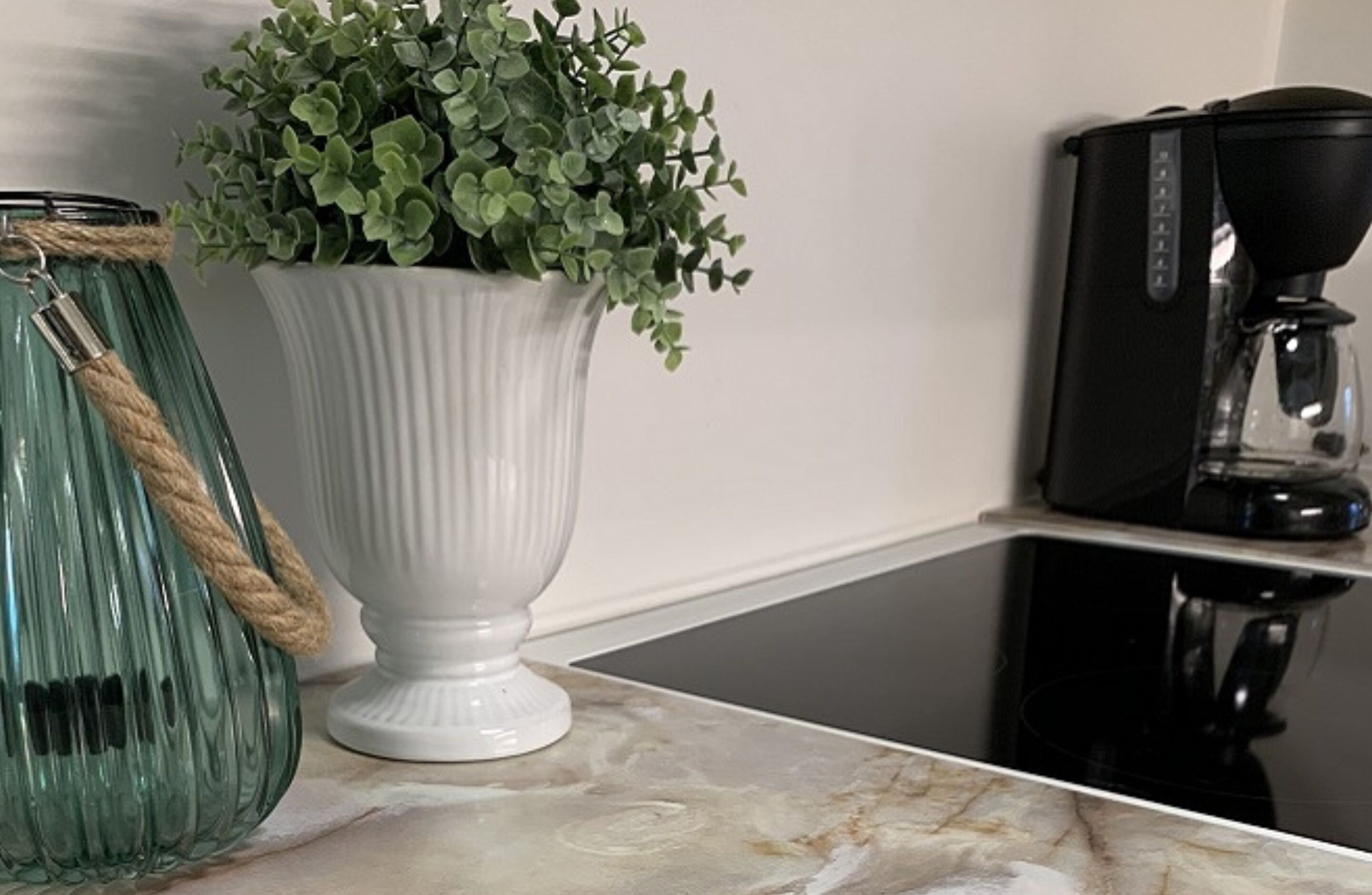 A kitchen counter with a white vase containing green foliage, a green glass jar with a rope, and a black coffee maker.
