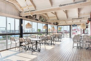 An indoor dining area with wooden tables and chairs, large windows, and hanging light fixtures. - First Camp Björkäng-Varberg (Tvaaker)