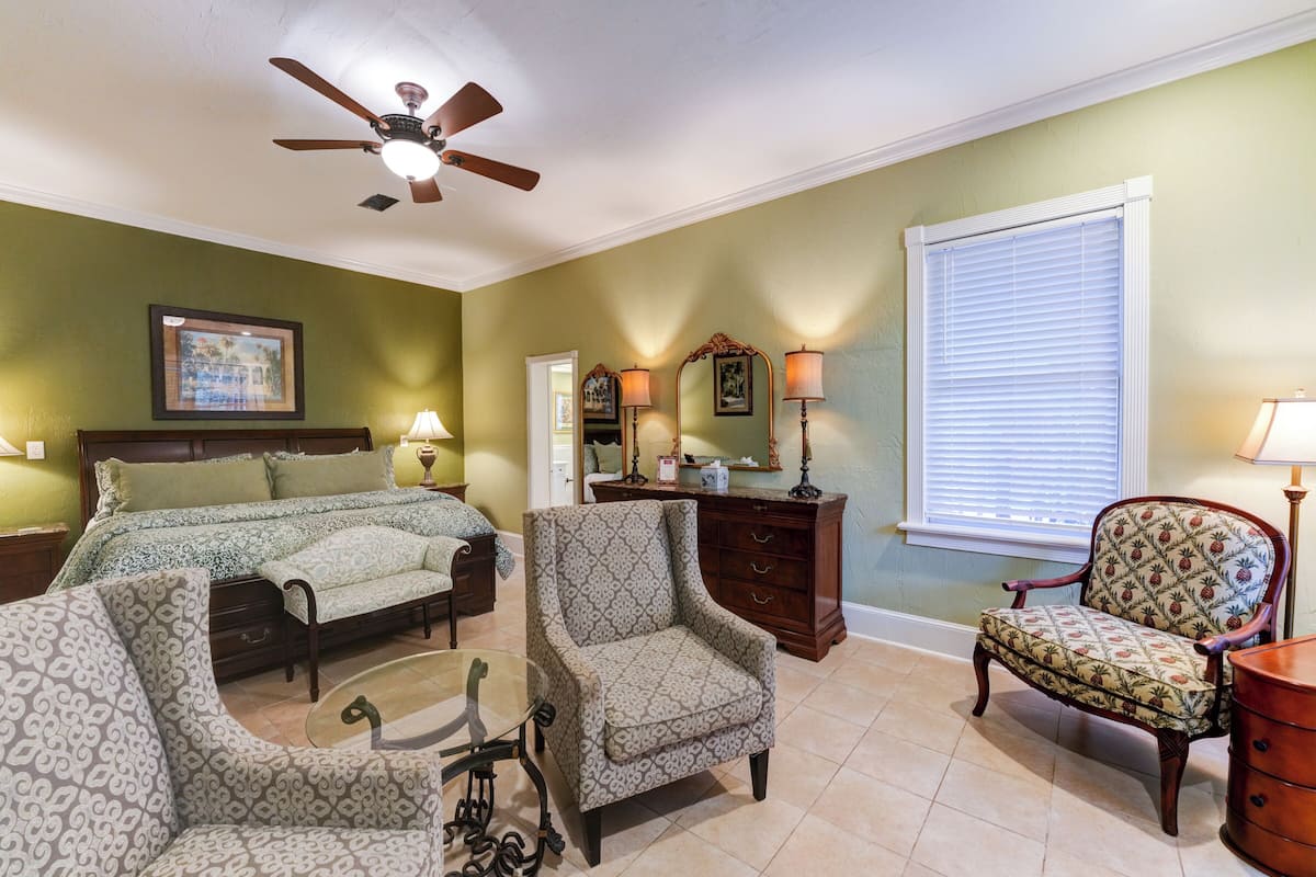 Une chambre à coucher comprenant un lit, deux fauteuils, une commode, un ventilateur de plafond et une fenêtre avec des stores.