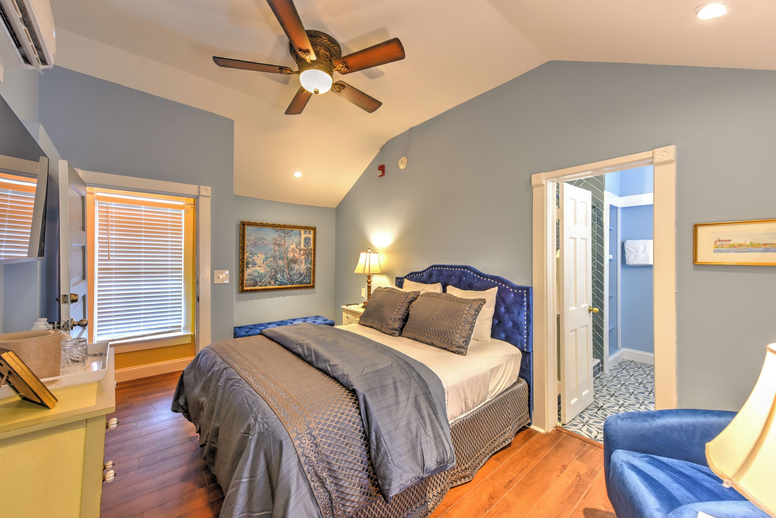 A bedroom with a bed, a ceiling fan, a nightstand, a lamp, a chair, and a door leading to another room.