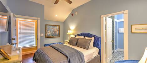 A bedroom with a bed, a ceiling fan, a nightstand, a lamp, a chair, and a door leading to another room.