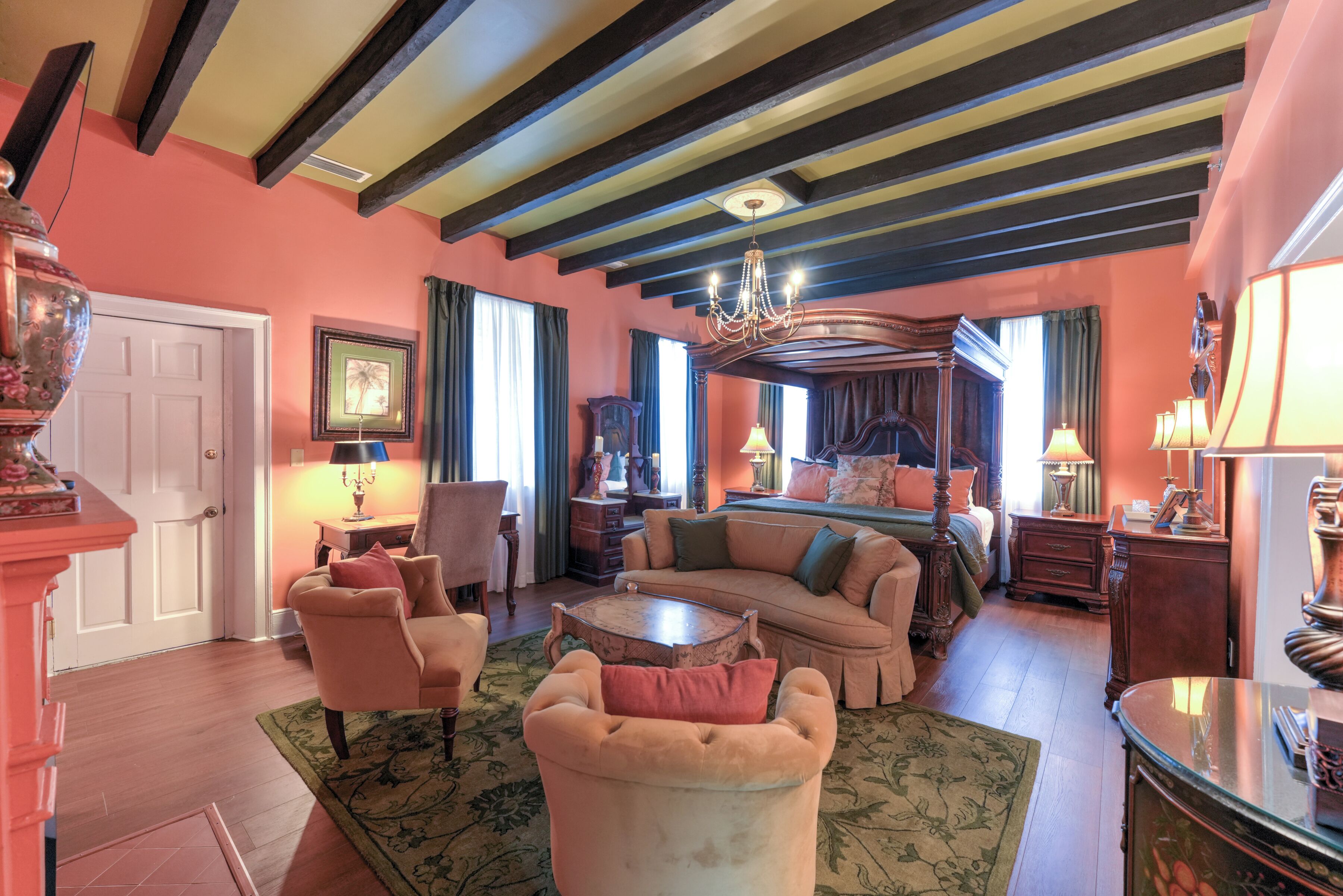 A room with a large bed, a sitting area with a sofa and chairs, a wooden coffee table, and a chandelier.