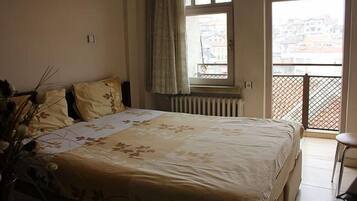 A bedroom with a bed, a radiator, a window with a view of buildings, and a balcony with a railing.