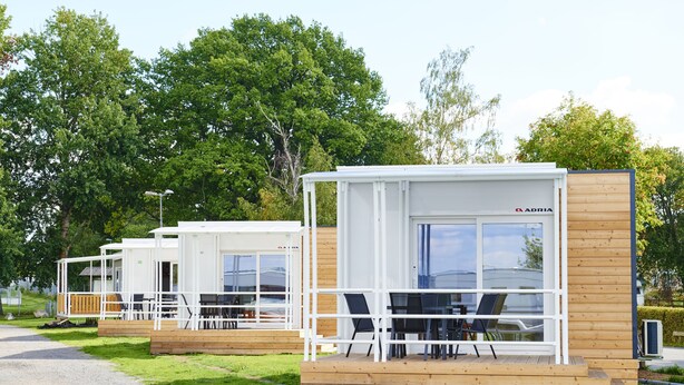 Une rangée de cabanes modernes, blanches et modulaires, dotées de terrasses en bois et d’espaces de détente en plein air, le tout entouré d’une végétation luxuriante.