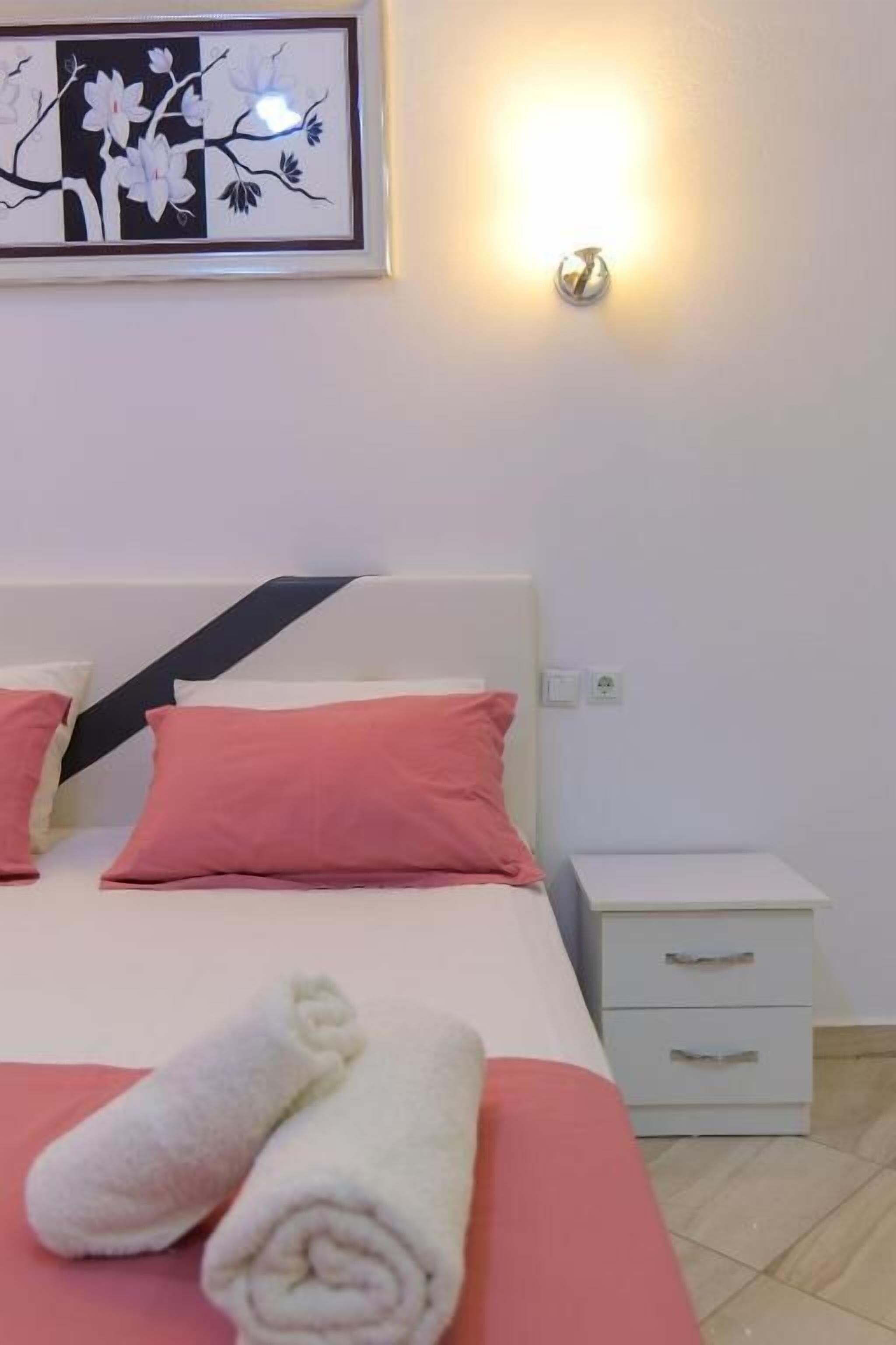 A neatly made bed with pink and white pillows, a framed artwork above it, and a bedside table with drawers.