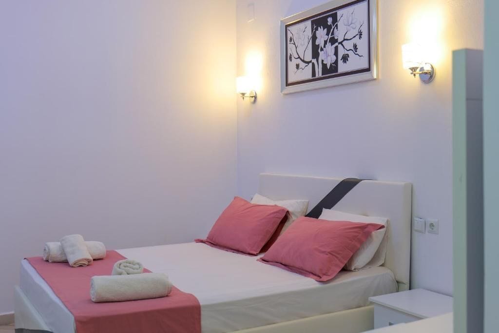 Ein ordentlich bezogenes Bett mit rosa und weißen Kissen, einer roten Decke und aufgerollten Handtüchern auf einer weißen Bettdecke.