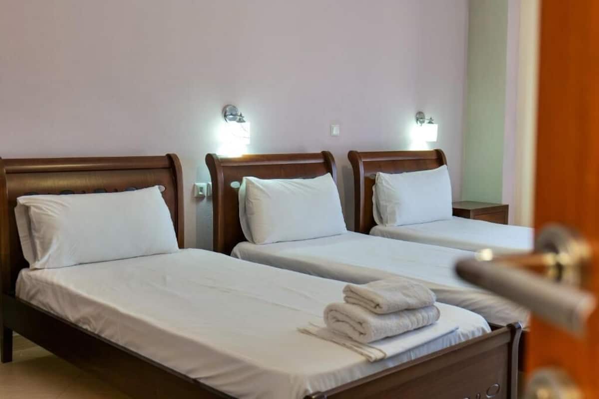 A hotel room with three single beds, each with white linens and pillows. There are wall-mounted light fixtures above each bed.