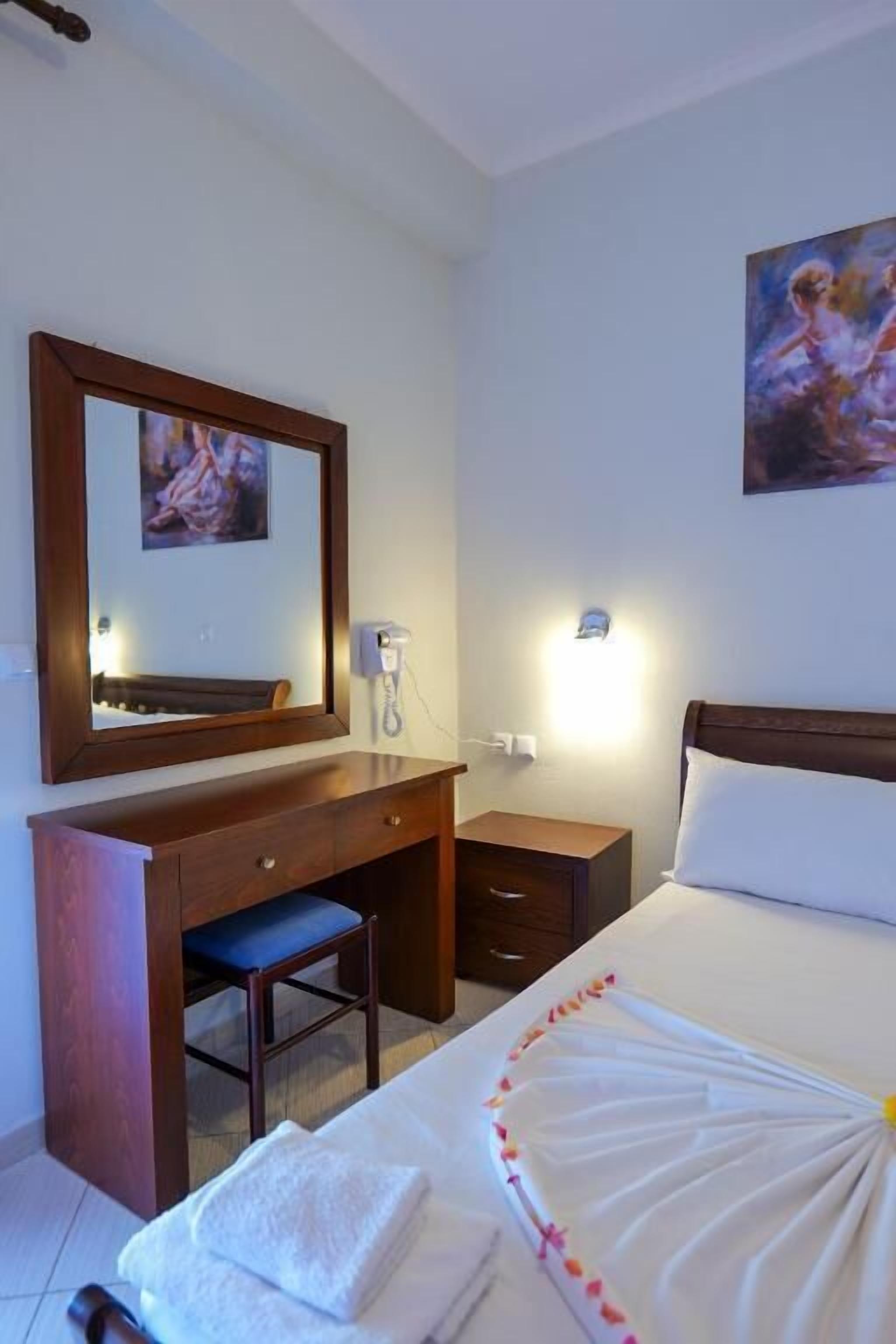 A hotel room with a bed, a desk with a chair, a bedside table, a nightstand, a mirror, and a wall-mounted light.