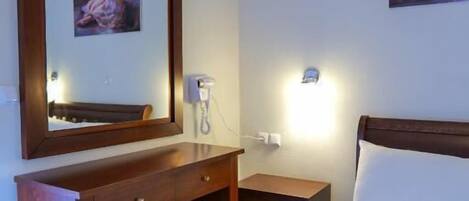 A hotel room with a bed, a desk with a chair, a bedside table, a nightstand, a mirror, and a wall-mounted light.