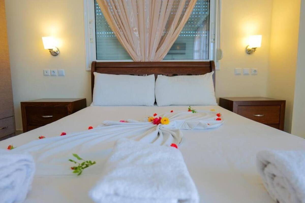 A neatly made bed with a floral arrangement and towels, a wooden headboard, bedside lamps, and a window with curtains.