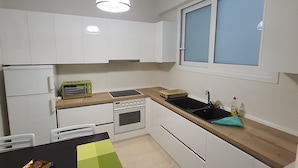 A compact kitchen with white cabinets, a built-in oven, a microwave, and a sink. There is a window with blinds, a trash bin, and a table with chairs. - Paradise City (Rethymno)